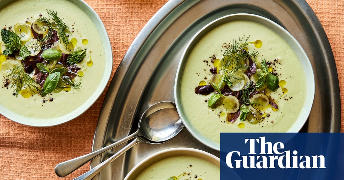 Three bowls of cream-coloured cold soup served in white and grey dishes, decorated with olives, fresh herbs and spices on an orange linen tablecloth.