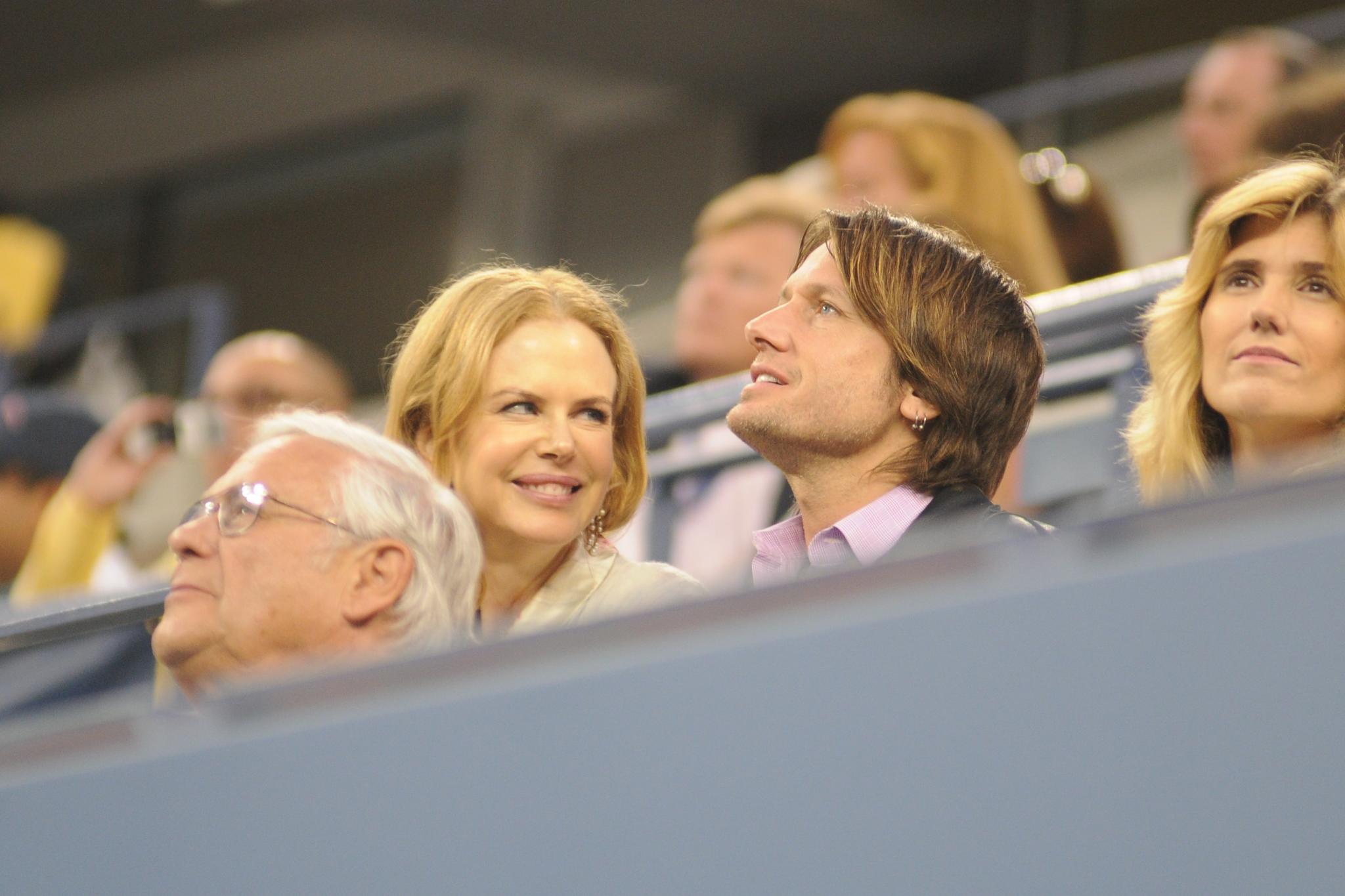 Nicole Kidman with reddish-blonde hair sits in stadium seating alongside an older man with glasses and a man with shoulder-length brown hair, watching an event.