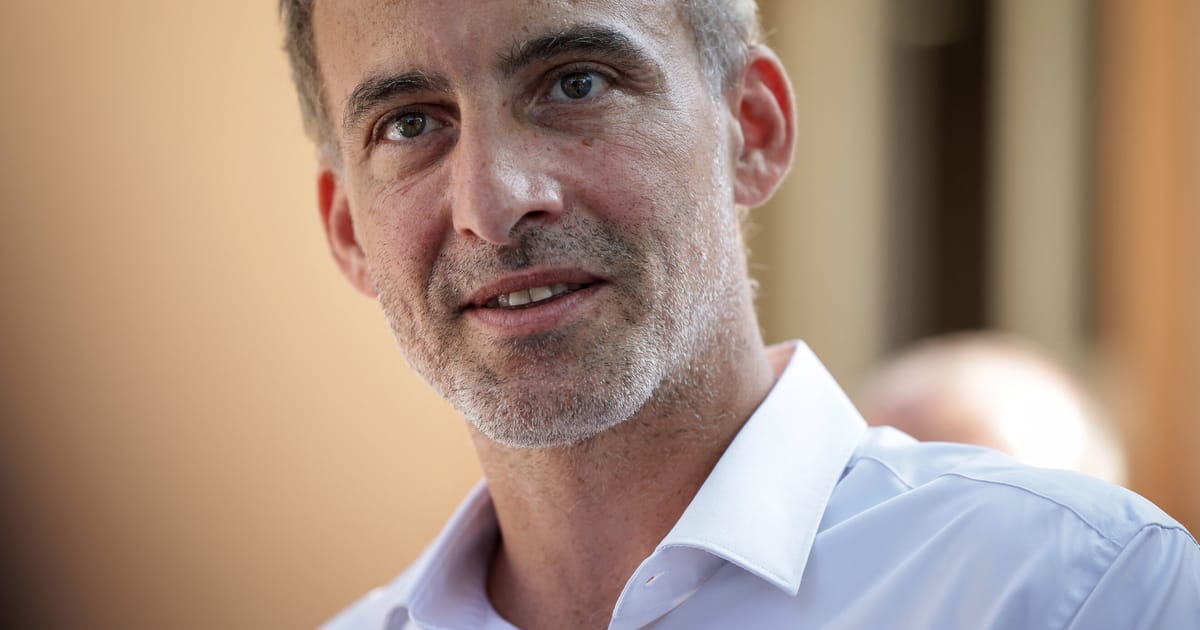 Retrato de un hombre de mediana edad con cabello gris y barba, vistiendo una camisa azul clara con cuello blanco, fotografiado contra un fondo desenfocado de tonos cálidos.