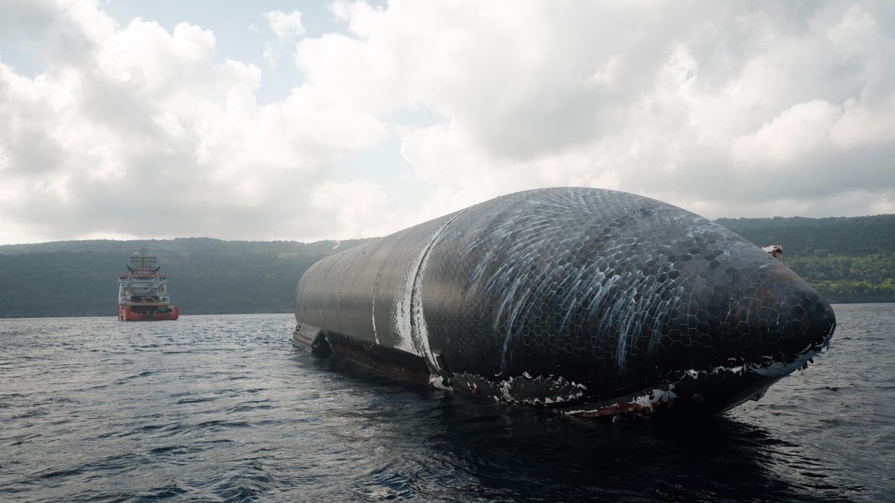 A large Starship vessel resting in ocean water with silver and black metallic structure, as a red and white tow ship approaches in the background.