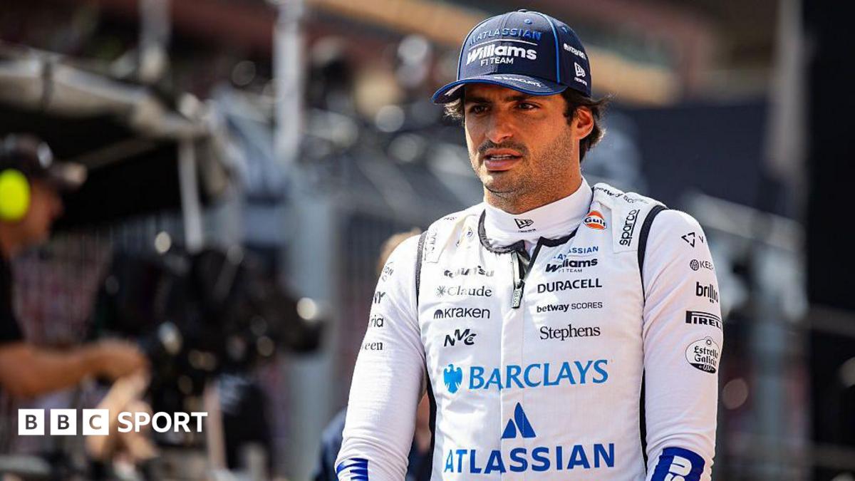 Un piloto de Fórmula 1 con traje de carreras blanco de Williams F1 Team y gorra azul está de pie en el garaje, mirando hacia la cámara.