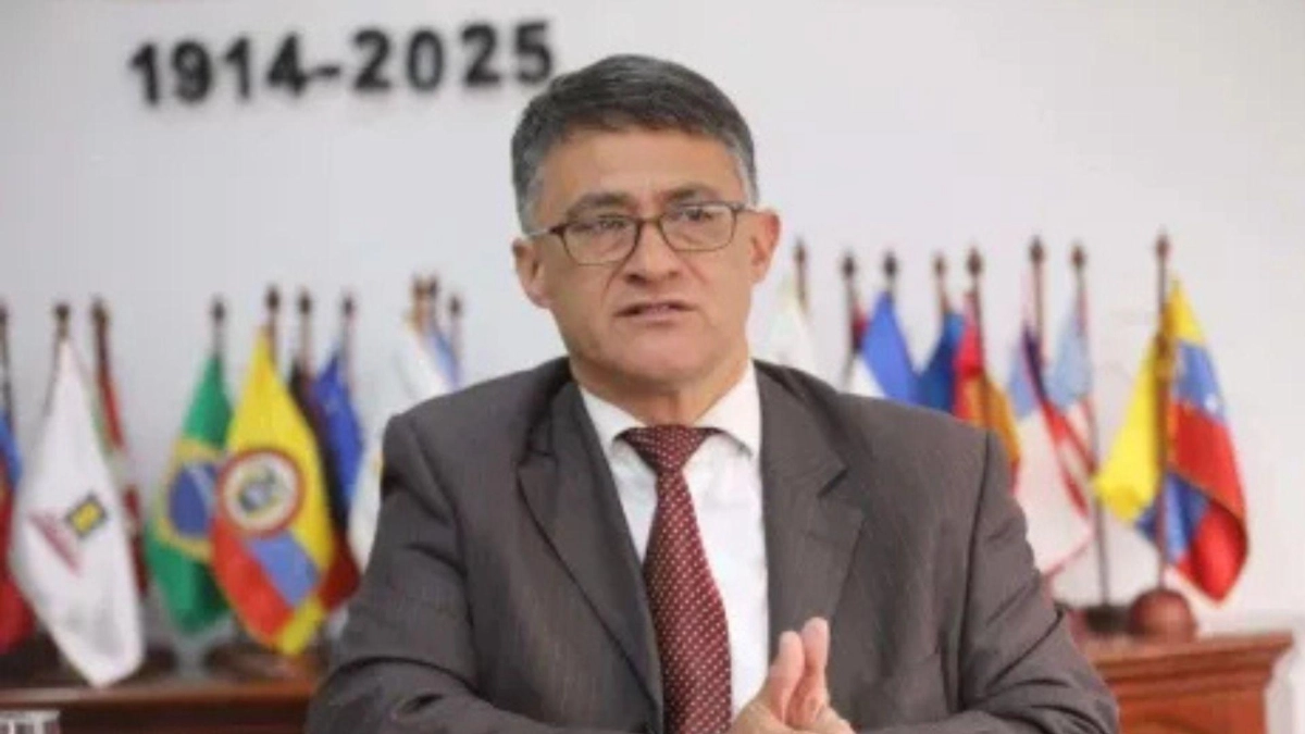 A middle-aged man wearing glasses and a dark grey suit with a red tie speaks at a podium, with flags of several countries and a banner reading "1914-2025" in the background.