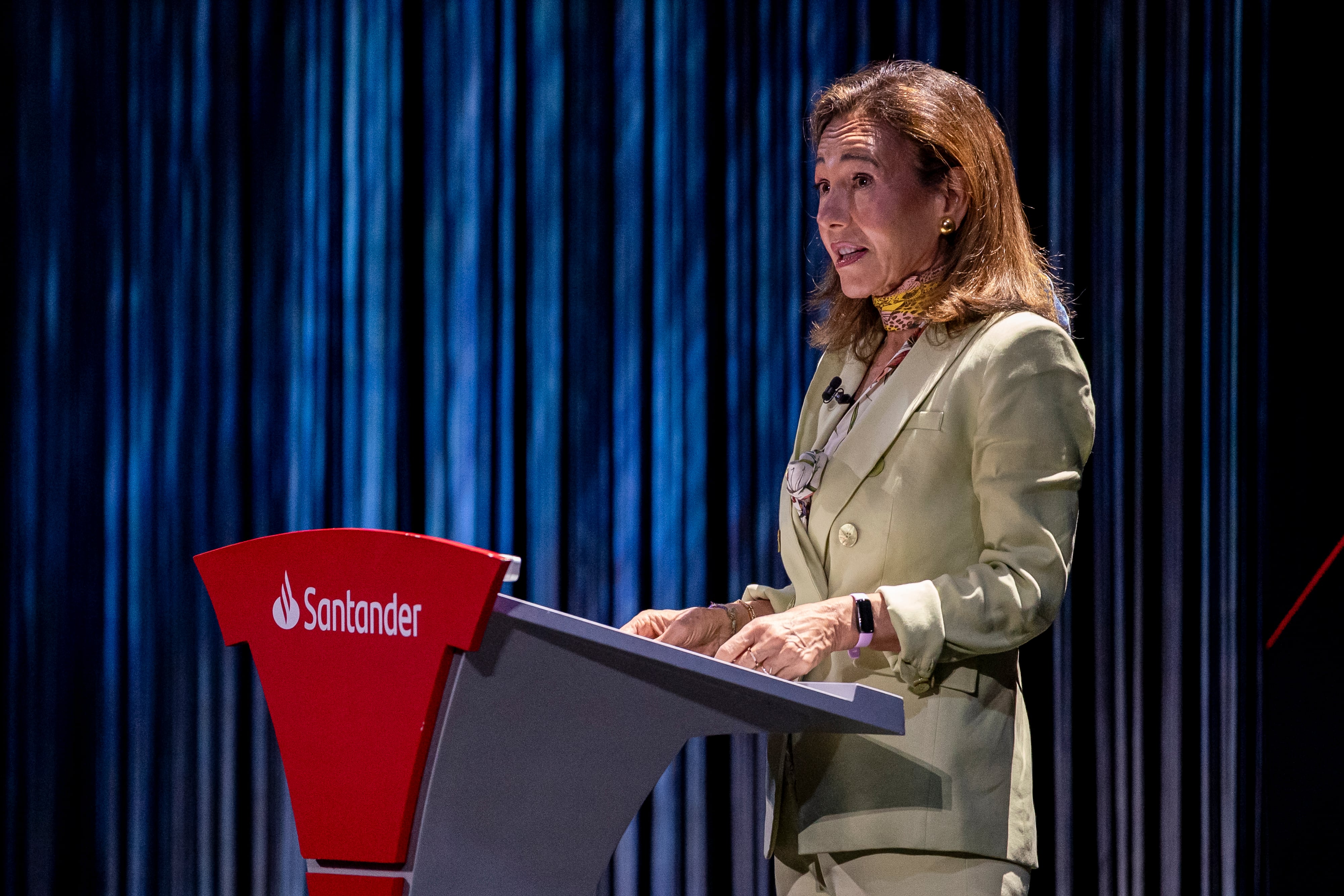 A woman in a light beige blazer stands at a podium with the Santander logo during a presentation against a blue curtain backdrop.