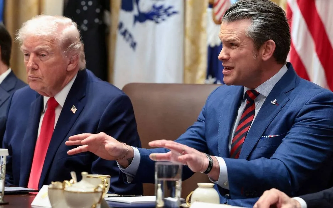 Two men in blue suits seated at a conference table, one wearing a red tie speaking and the other listening, with U.S. flags in the background.