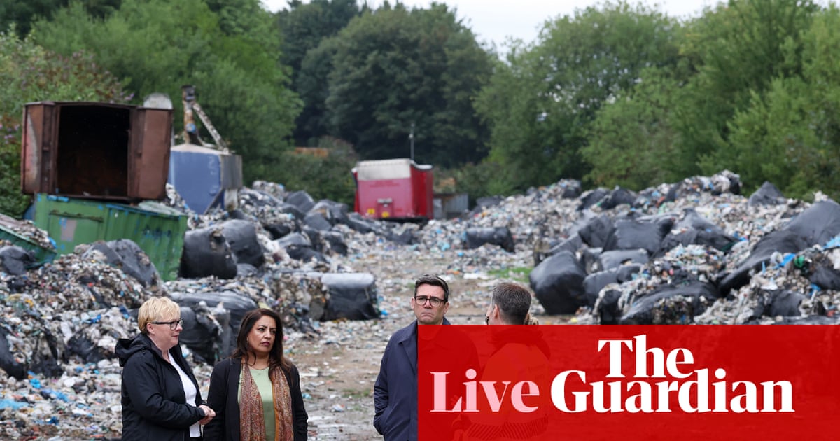 Three officials walk through a sprawling landfill site filled with piles of waste, rubble, and black bags, with equipment and shipping containers visible in the background.