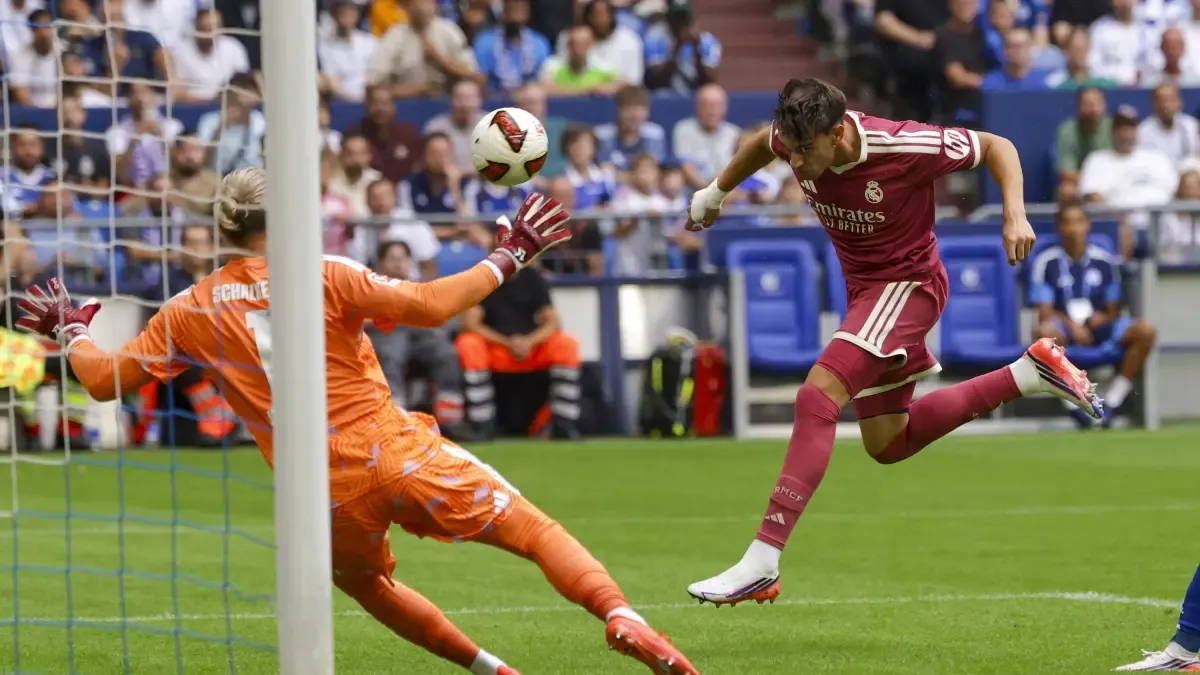 A Real Madrid player in burgundy kit attempts to shoot the ball while a goalkeeper in orange tries to block it.