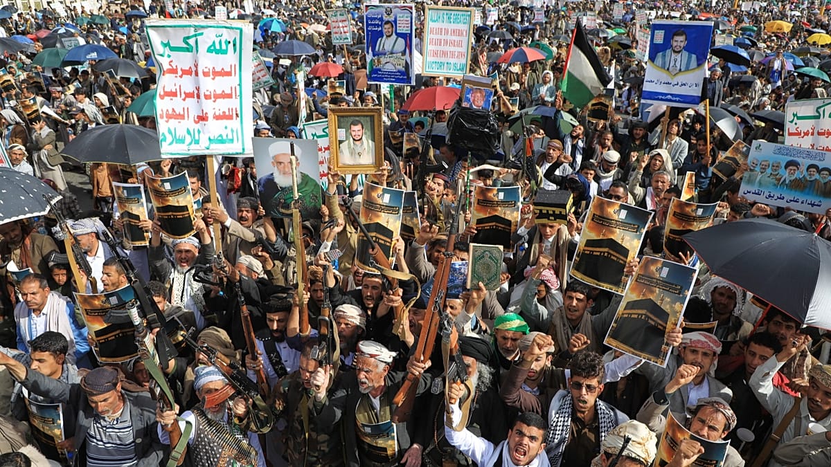 Una multitud de manifestantes se reúne en una plaza, sosteniendo armas, carteles con texto en árabe y retratos de personas, bajo paraguas y con banderas palestinas visibles.