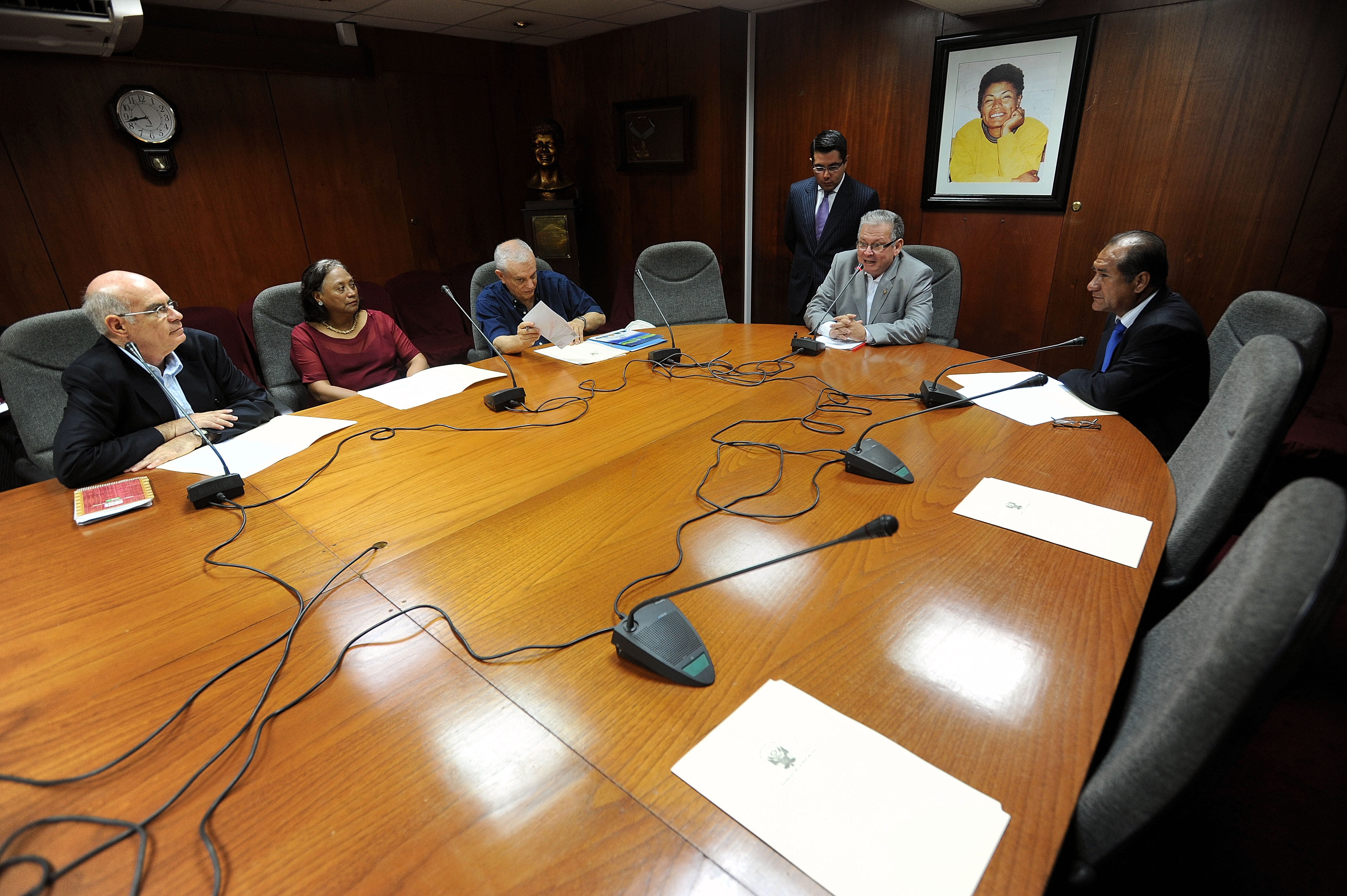 A group of people gather around a wooden table in a wood-panelled conference room, with microphones placed in front of several attendees.