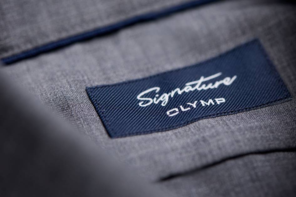 A close-up of a dark blue fabric label with the "Signature OLYMP" logo embroidered in white on the grey fabric of a garment.