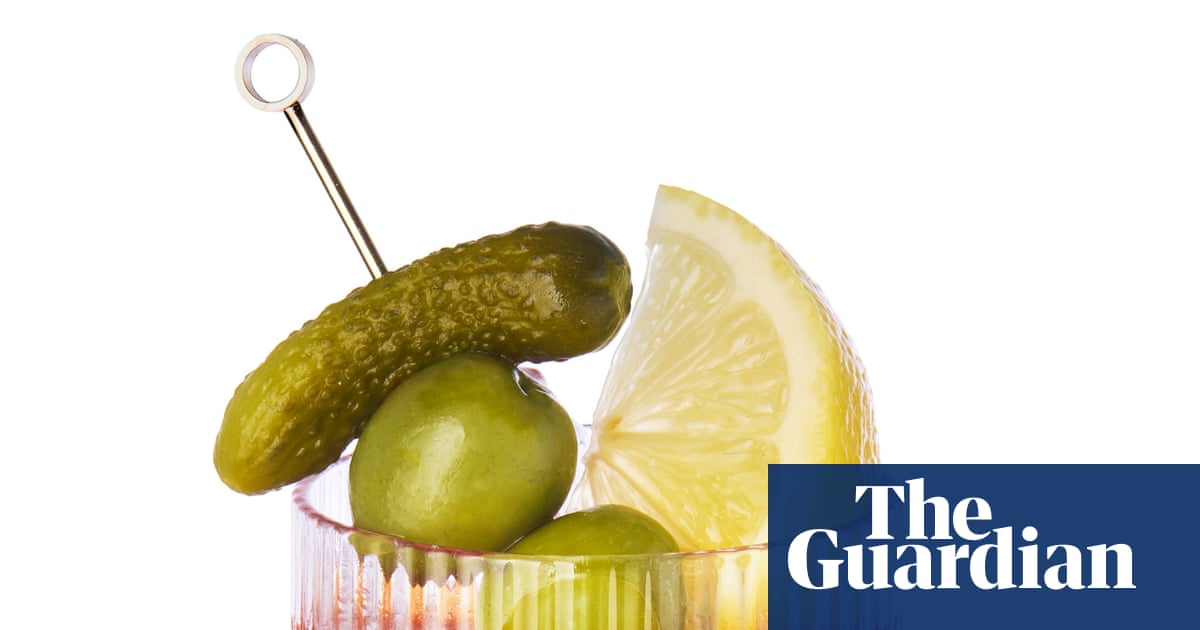 Un vaso con una bebida que contiene un pepinillo ensartado en una brocheta de metal, una aceituna verde, y una rodaja de limón amarillo.