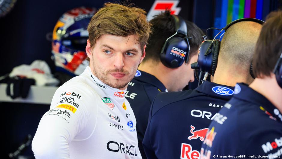 A Formula 1 driver in a white Red Bull uniform stands in the garage, talking with team members wearing dark blue uniforms.